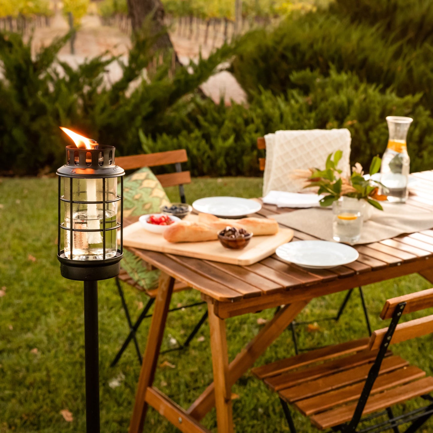 Black outdoor torch with flame, set on grass near wooden table with bread, plates, and glasses.
