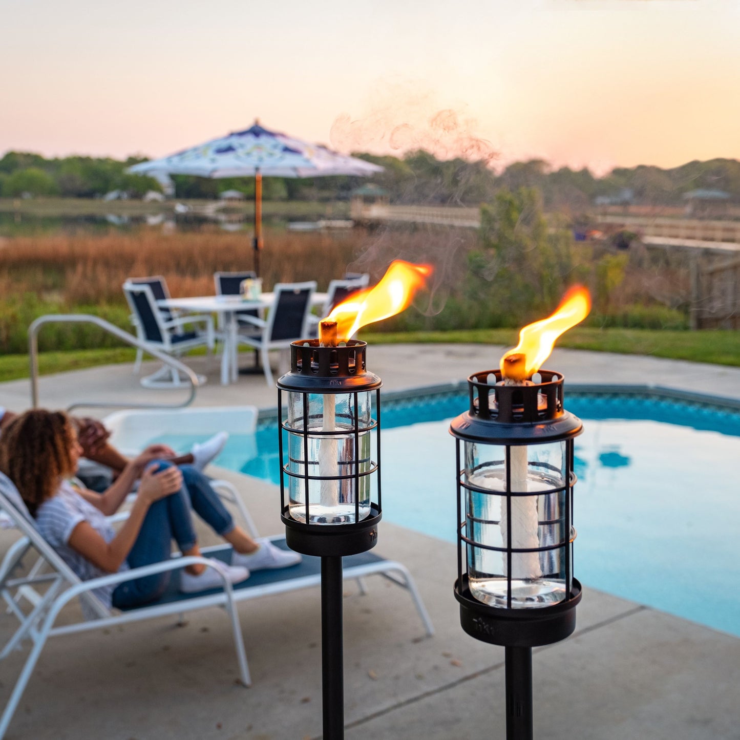 Black outdoor torches with glass cages and bright flames, near a poolside setting.