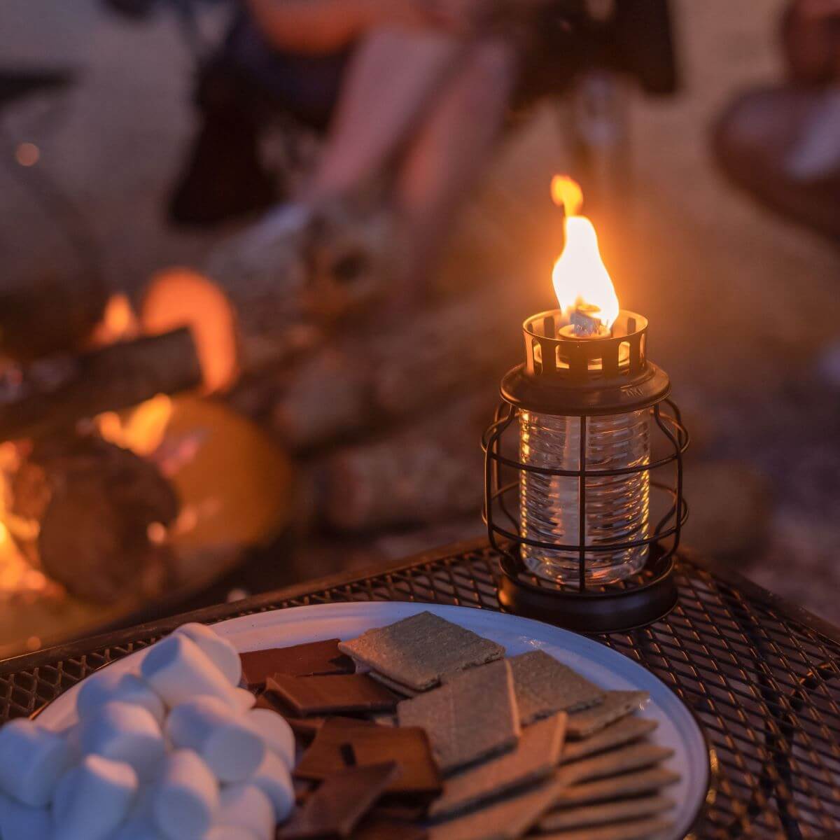 Black metal lantern with flame, beside plate of graham crackers and marshmallows.