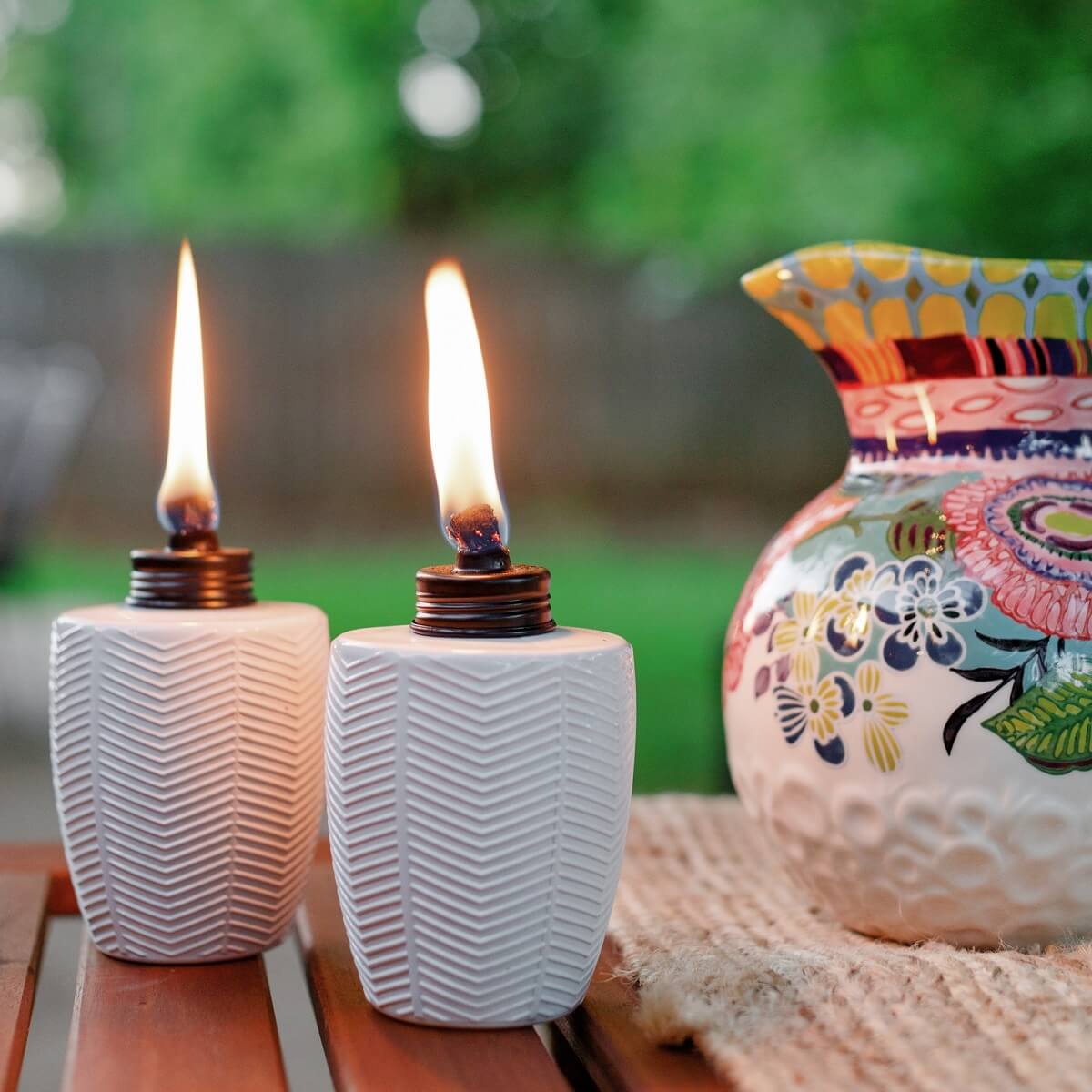 Two white ceramic oil lamps with flame, beside a colorful floral vase.