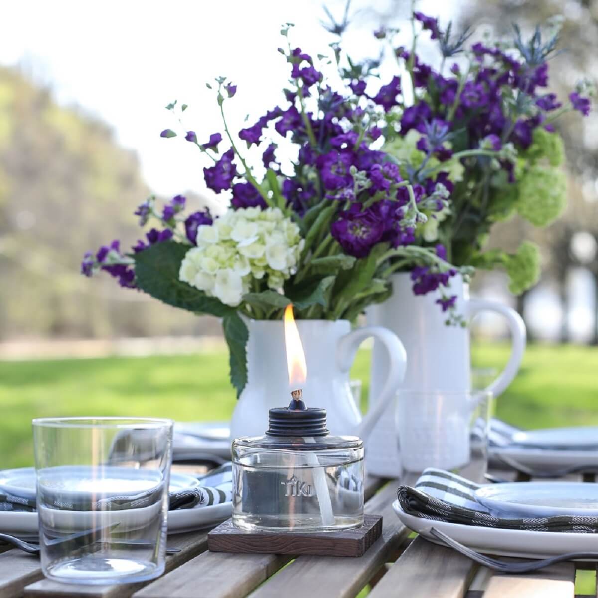 Clear glass TIKI oil lamp with flame, set on table with flowers and plates.