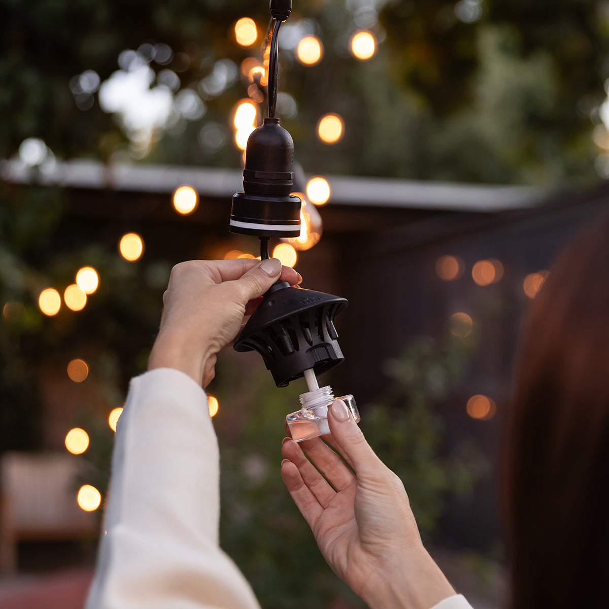 Outdoor insect repellent diffuser being refilled, black hanging device with clear refill bottle, warm lights in background.