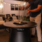 Black outdoor fire pit table with spiral safety cage, set on patio with string lights.