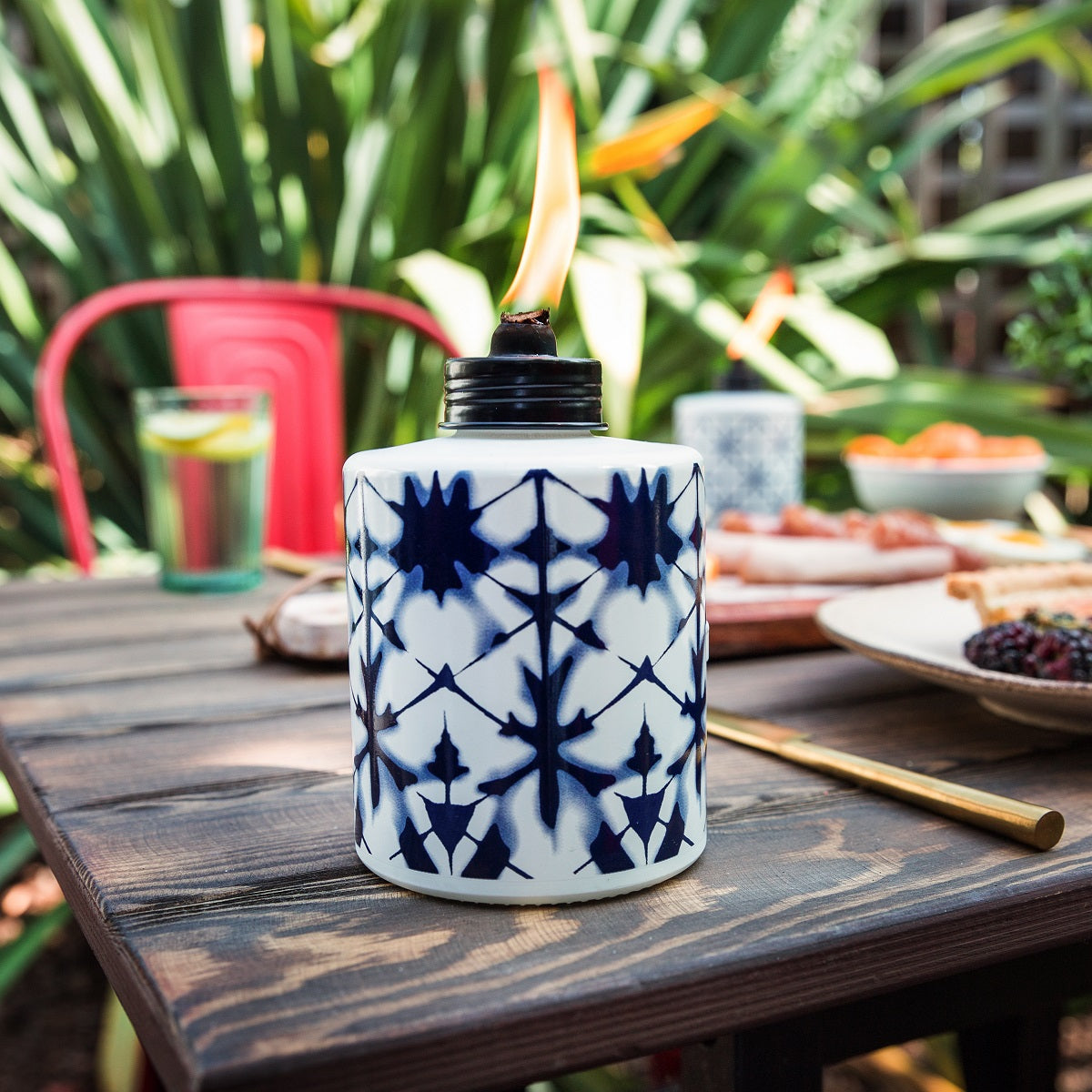 White ceramic tiki torch with blue geometric pattern, lit flame, outdoor setting.