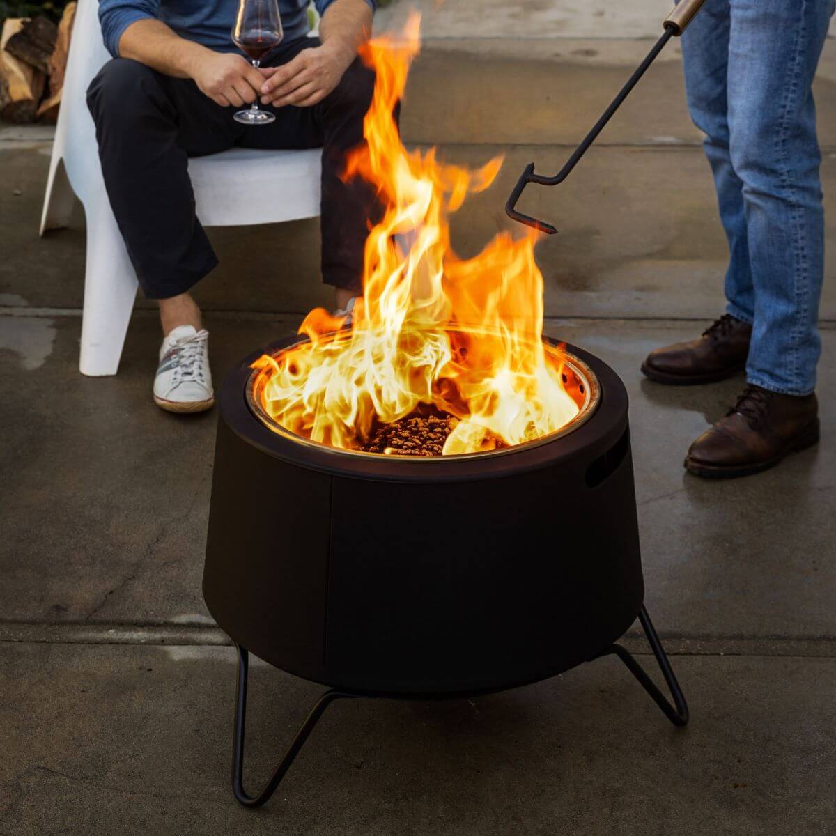 Black fire pit with flames, used outdoors with people nearby.