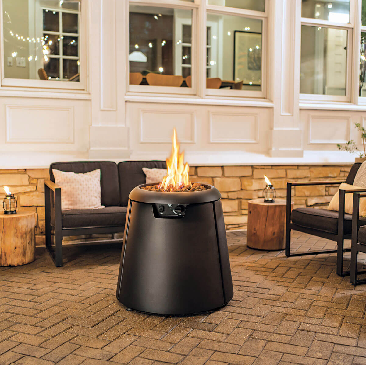 Black fire pit with flames, surrounded by outdoor furniture and brick flooring.