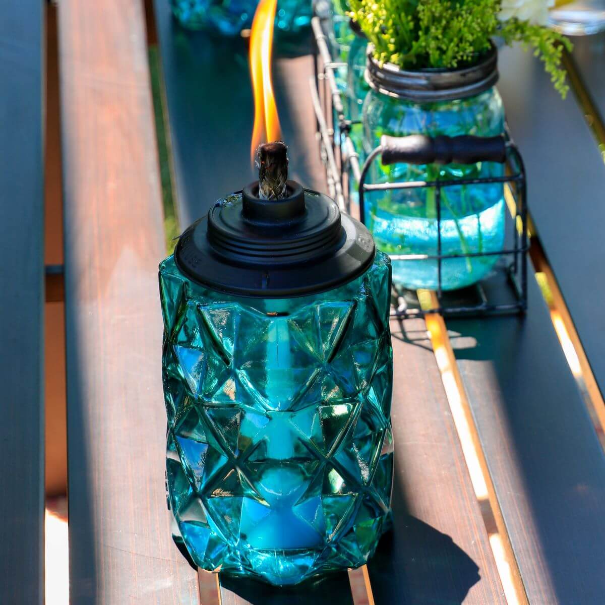 Blue glass outdoor tiki torch with geometric pattern, black metal top, and visible flame, placed on a table.
