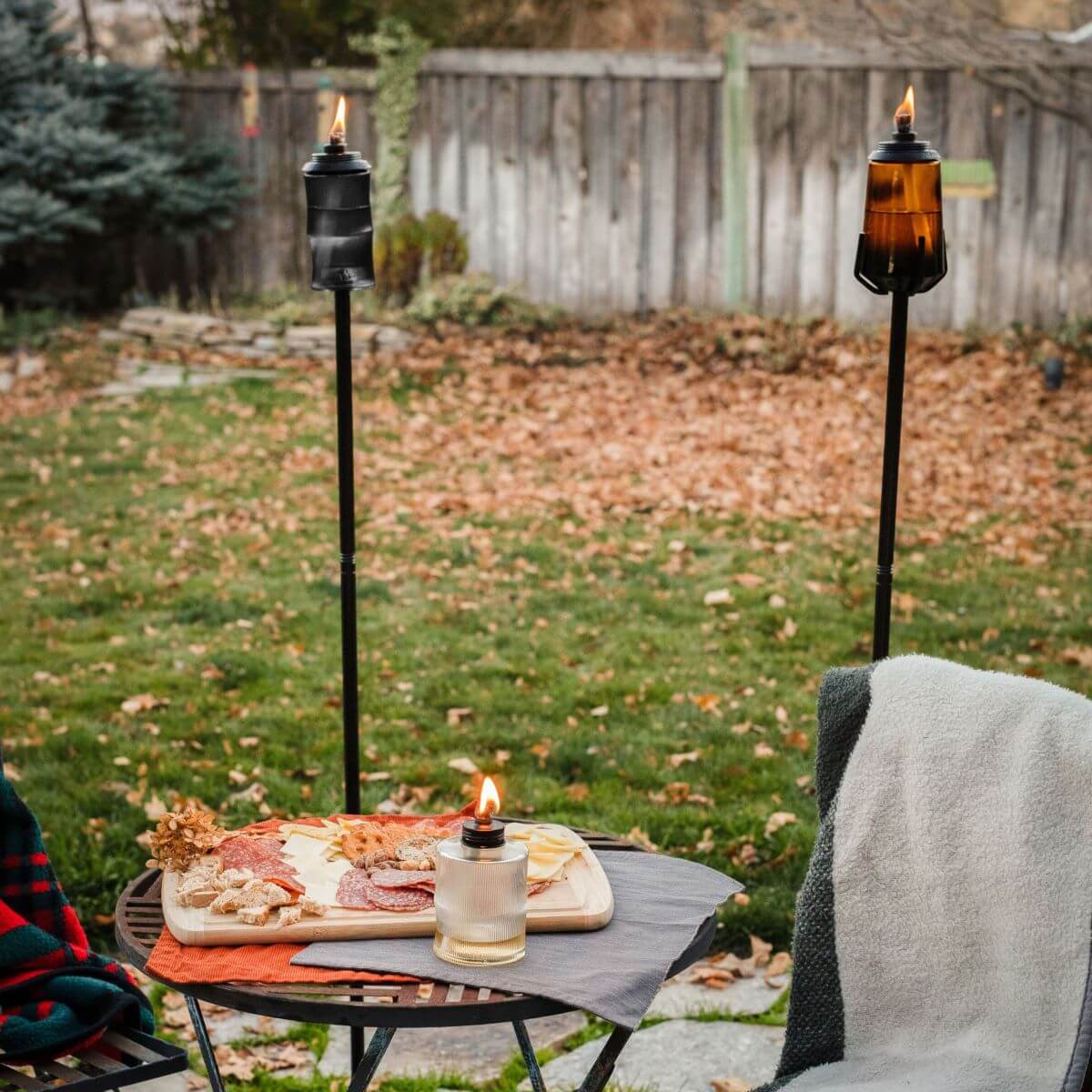 Tall black outdoor torches with flames, set on grassy yard with picnic table and blanket.