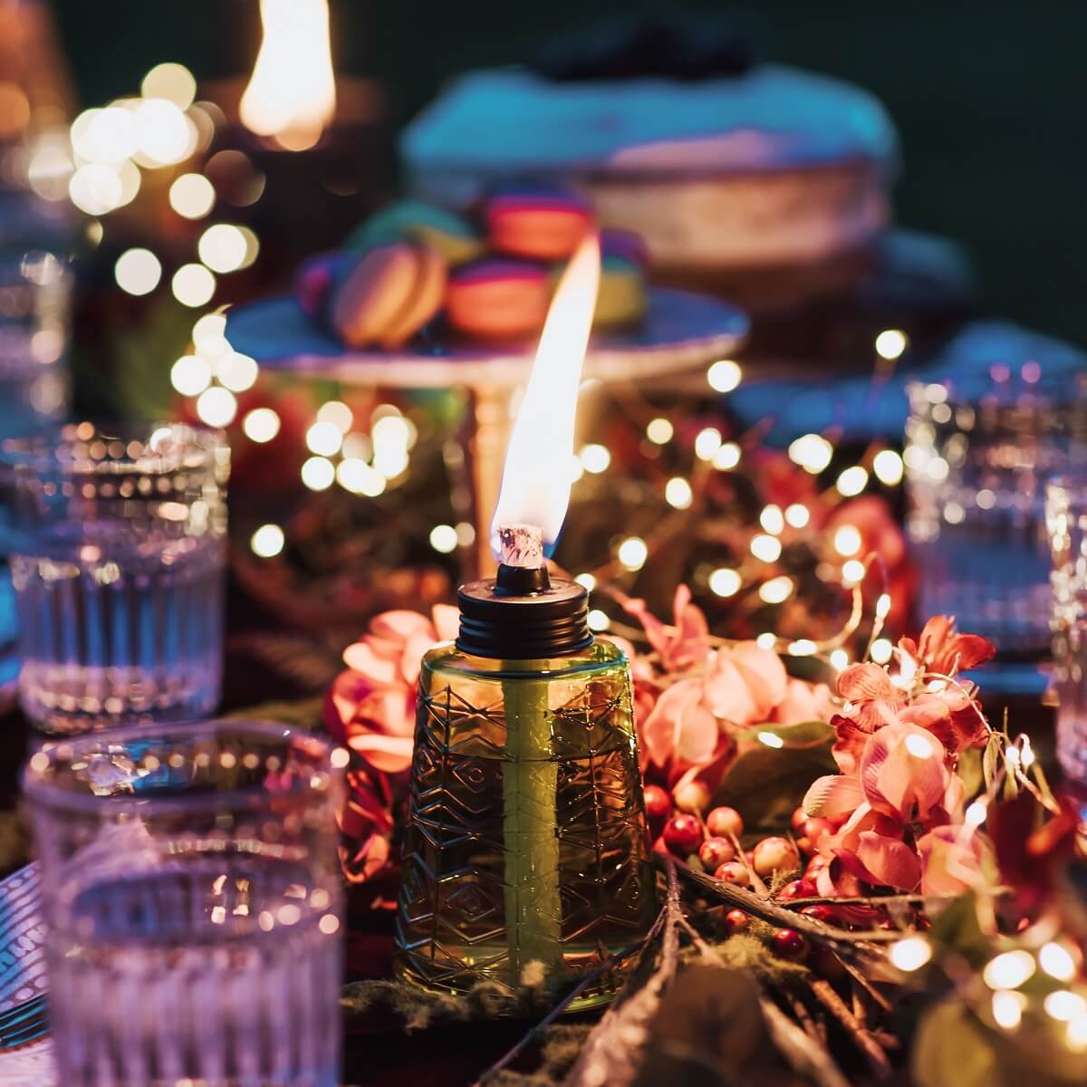 Green glass oil lamp with flame, surrounded by festive lights and flowers.