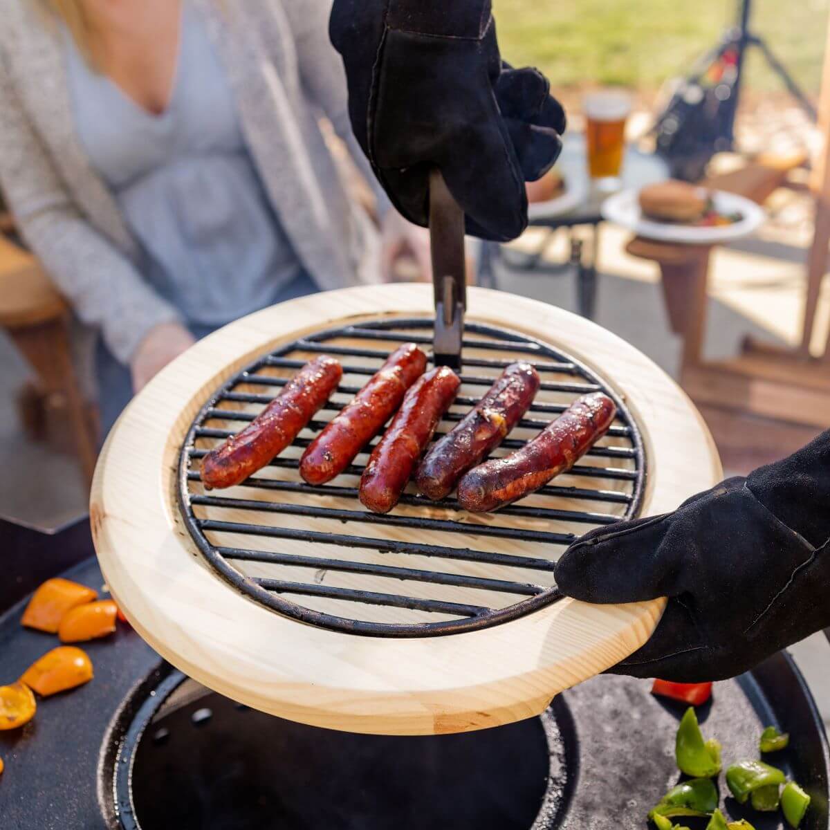 Wooden tray with black grill holding sausages, hands in black gloves cooking outdoors.