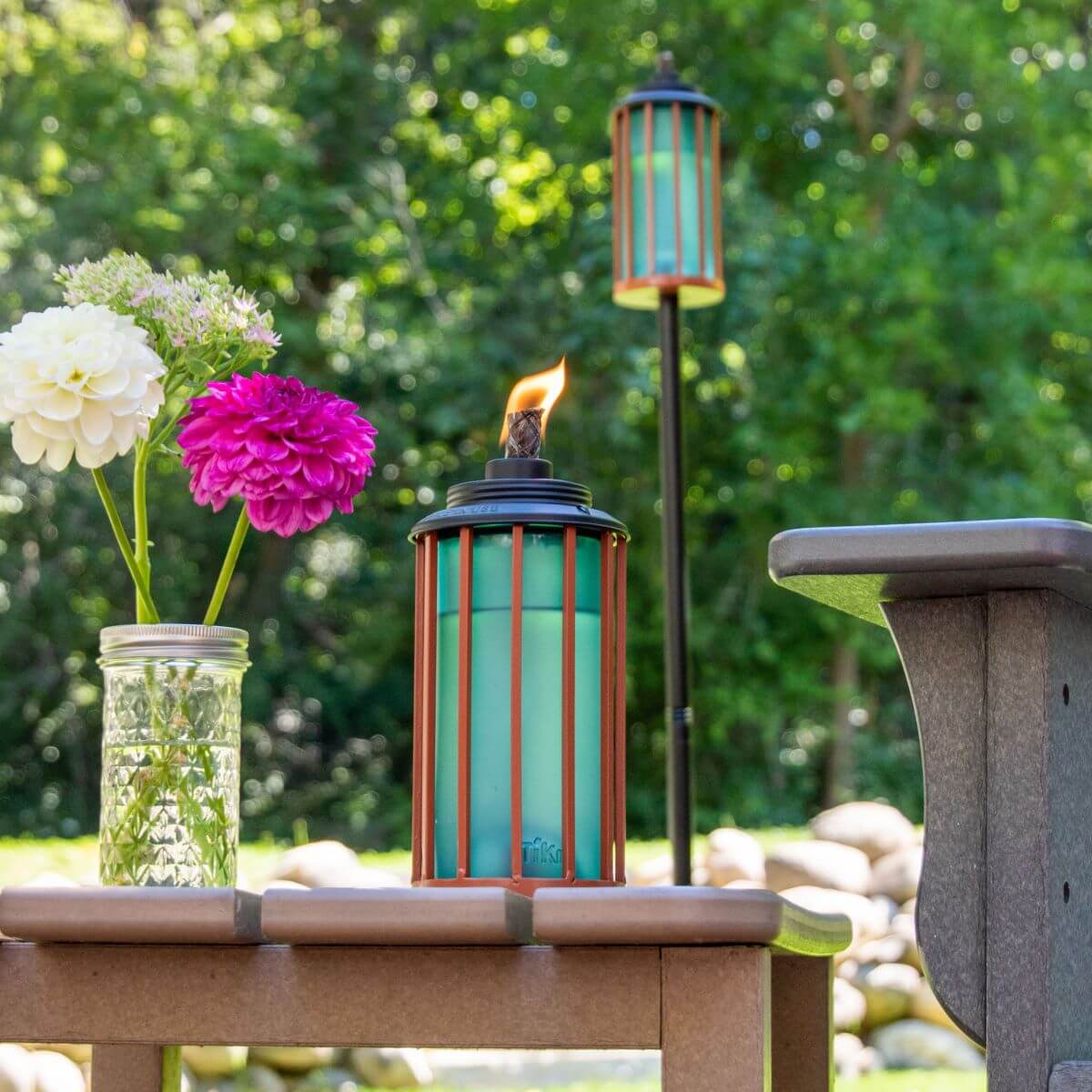 Tiki torch with teal and red cage, lit flame, beside flowers in mason jar.