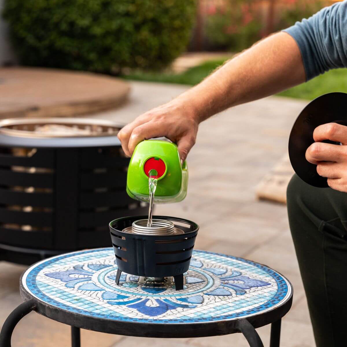Green fuel container pouring liquid into blue fire pit burner on mosaic table.
